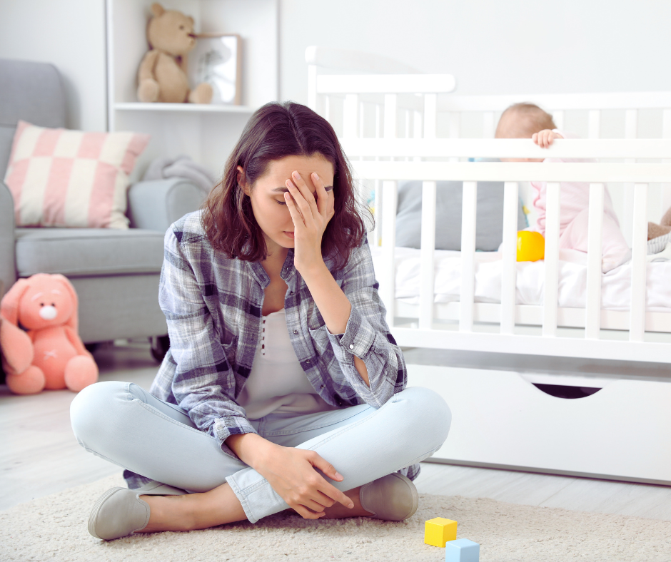 Woman with postpartum depression sitting next to her baby's crib with her hand over her face.